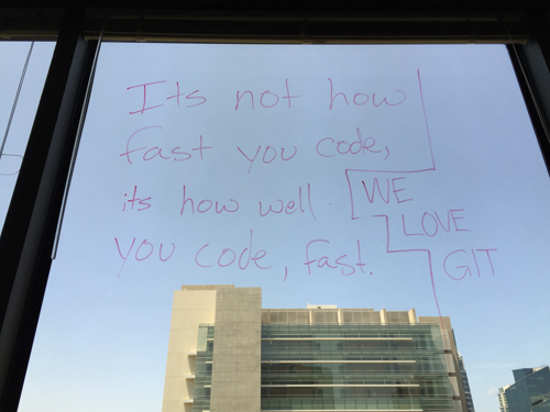 whiteboard marker writing on the inside of a skyscraper window with the quote "It's not how fast you code, it's how well you code, fast" and "We love Git"