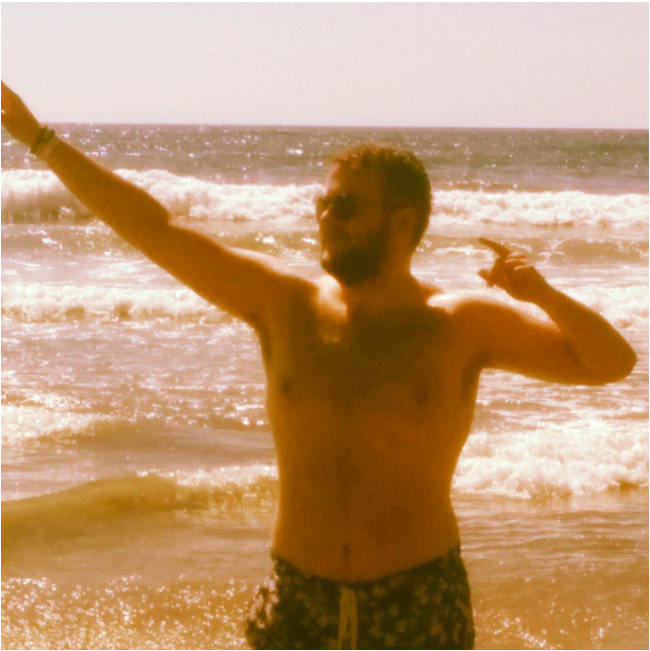 a shirtless bearded man at the beach, flexing and pointing off camera
